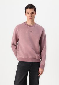 Helles rosa Sweatshirt aus weichem Material; runder Ausschnitt, gerippte Ärmelbündchen und Saum; mit schwarzem Textlogo auf der Brust; kombiniert mit dunklen Jeans.