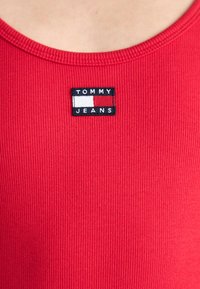 Raudona ripsuota marškinėlių su juodu stačiakampiu ženkleliu, kuriame užrašyta "TOMMY JEANS" ir mažas baltas bei raudonas logotipas. Slydus paviršius.