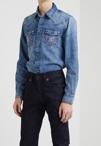 Denimskjorta i ljusblått med två bröstfickor. Mörka jeans med röda knapputskärningar. Har en västernstil krage och kontrasterande sömmar.