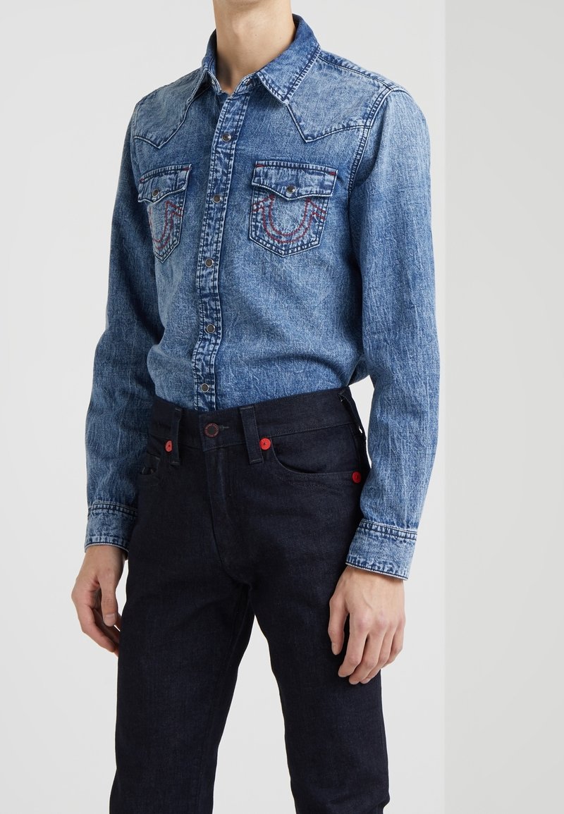 Denimskjorta i ljusblått med två bröstfickor. Mörka jeans med röda knapputskärningar. Har en västernstil krage och kontrasterande sömmar.