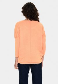 Orangenes, leichtgewichtiges Strickoberteil mit Dreiviertelärmeln, lockerer Passform, abgerundetem Saum und einer mittigen Rückennaht. Getragen über dunkelblauen Jeans.