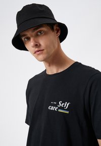 Czarna bawełniana koszulka z białym napisem "Self care" i "est. 1988" z przodu. Noszona z czarną kapeluszem typu bucket. Gładka faktura, swobodny fason.