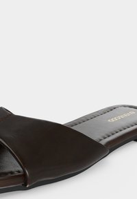 Sandale en cuir marron foncé à bride large avec logo doré sur une semelle rembourrée, présentée sur fond blanc.