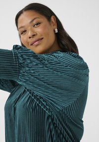 Teal-rippierter Stoffoberteil mit strukturierten vertikalen und diagonalen Falten. Mit gerafften Ärmeln und subtiler Glanz. Silberne Ohrringe runden den Look ab.