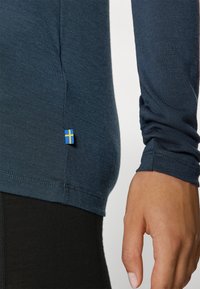 Long-sleeve navy shirt made of textured fabric, fitted design, featuring a small blue and yellow flag detail at the hem.