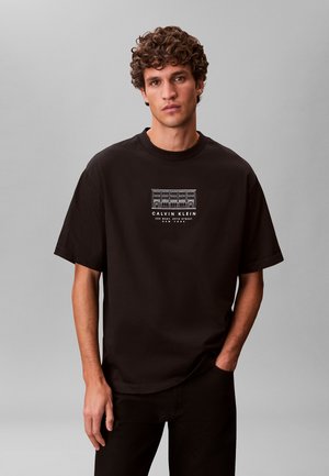 Junger Mann mit lockigem Haar trägt ein schwarzes Calvin Klein T-Shirt mit einem weißen architektonischen Gebäudegrafik und Text auf der Brust.