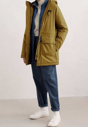 Persona che indossa un parka color senape sopra una giacca di jeans blu, un maglione grigio, pantaloni blu risvoltati e scarpe da ginnastica bianche alte.