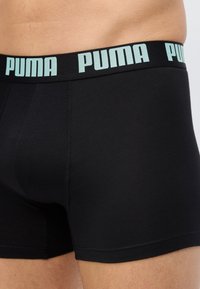 Μαύρα ανδρικά εσώρουχα μποξέρ με φαρδιά ζώνη μέσης που φέρει επαναλαμβανόμενα λογότυπα "PUMA" σε ανοιχτό μπλε, φορεμένα από άτομο που δείχνει τη μέση και το άνω μέρος των μηρών.