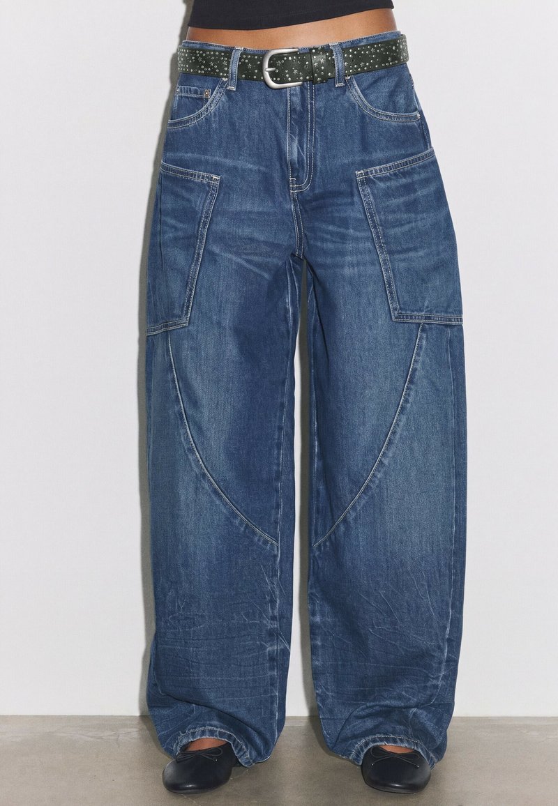 Jean bleu à jambes larges avec grandes poches latérales, porté avec une ceinture noire cloutée et des chaussures noires sur un sol en béton.