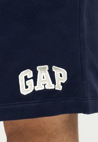 Tissu bleu marine avec les lettres "GAP" brodées en blanc près de l'ourlet au-dessus de la jambe visible d'une personne.
