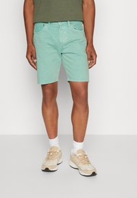 Pantalones cortos de mezclilla en un azul claro, con un corte clásico, que presentan bolsillos frontales, pasadores para cinturón y una cremallera. Combinados con zapatillas beige y calcetines blancos.