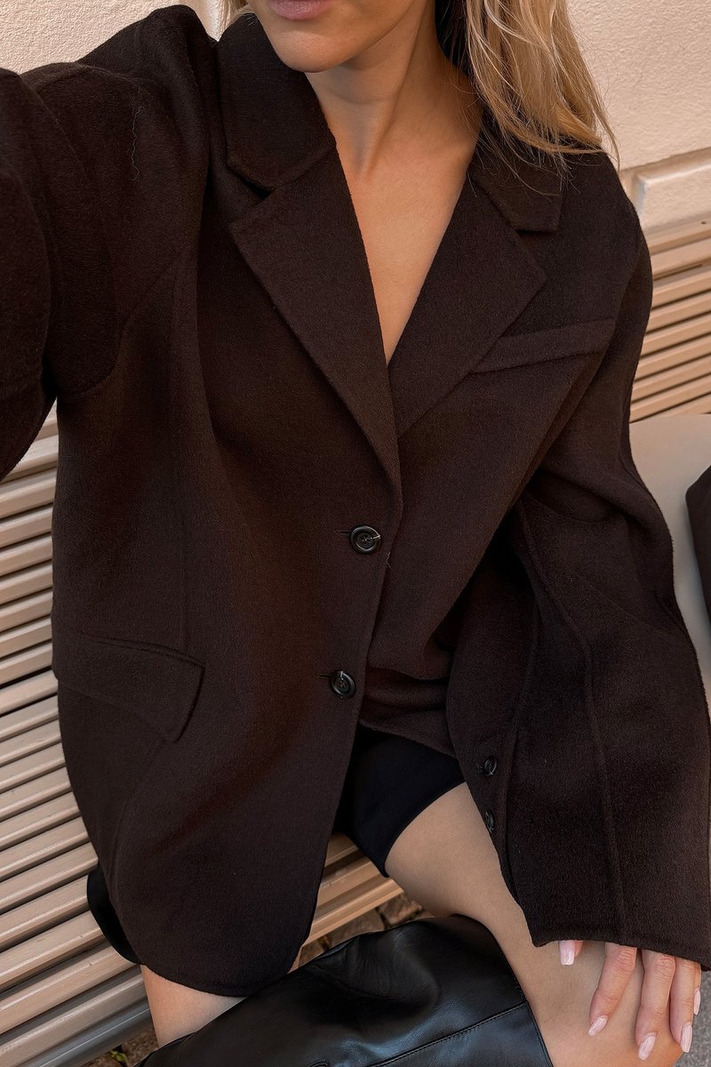 Temno rjava blazer iz volnene mešanice, ki se ponaša s krojenjem, zarezanimi reverji, dvema gumboma in stranskima žepoma, v kombinaciji s črnim oblačilom.