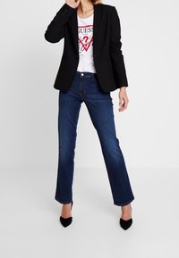 Blazer nero su una t-shirt bianca con grafica, abbinato a jeans a zampa d'elefante blu scuro e tacchi a punta neri. Texture morbide e vestibilità sartoriale.