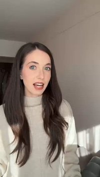 Pessoa com cabelo longo e escuro, vestindo um suéter bege claro, de pé contra uma parede neutra, engajando-se numa conversa com uma expressão clara.