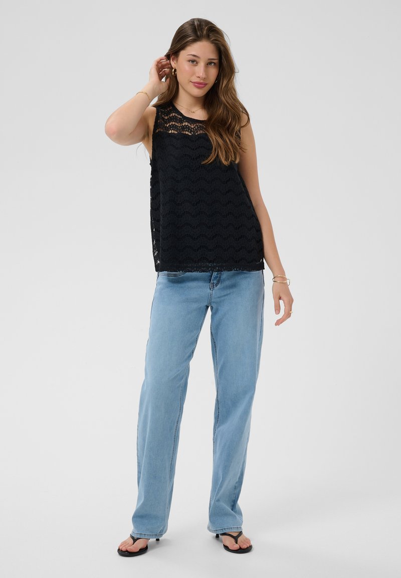 Jeune femme aux longs cheveux portant un top en dentelle noire sans manches, un jean droit bleu clair et des sandales noires, debout devant un fond uni.