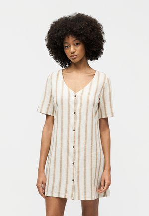 Γυναίκα με σγουρά μαλλιά φοράει κοντομάνικο, κουμπωτό φόρεμα με V λαιμόκοψη και κάθετες μπεζ και λευκές ρίγες, στέκεται μπροστά σε ένα απλό λευκό φόντο.