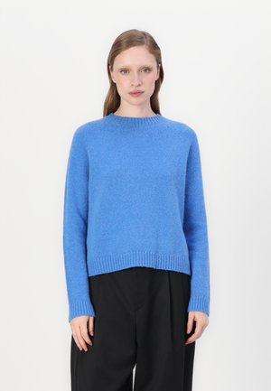 Blue, textured sweater with long sleeves and a rounded neckline. Ribbed hem and cuffs. Worn over black, loose-fitting pants.