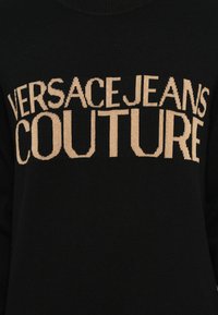 Melna adīta džemperis ar tekstu "VERSACEJEANS COUTURE", kas izcili redzams bežā krāsā uz priekšpuses. Klasiska apaļa kakla dizains.