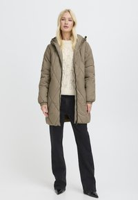 Gepolsterter, olivgrüner Daunenmantel mit Kapuze, ausgestattet mit einem Reißverschluss und seitlichen Taschen, getragen über einem cremefarbenen, grob gestrickten Pullover und schwarzen Jeans.