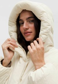 Svetlo béžová páperová bunda s veľkou kapucňou, so zipsovým uzatváraním a prešívanou textúrou. Ruky držia golier, pričom ukazujú jemnú látku.