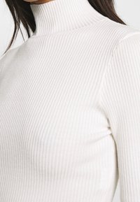 Haut à col roulé côtelé en matière douce blanc cassé. Il présente un col haut, une coupe ajustée et une surface texturée pour un intérêt visuel.