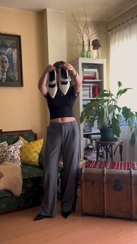 Valge ja musta kärgstruktuuriga terava nina sandaalid, millel on pahkluuvöö, neid hoiab musta särgiga ja hallide pükstega inimene.