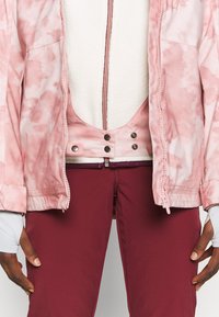 Rosa Batikjacke mit Reißverschluss und Druckknöpfen, über einem weißen Fleece-Innenfutter, kombiniert mit bordeauxroten Hosen.