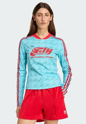 Ljusblå långärmad skjorta med röda detaljer och texten "Fly Tanzbar", i kombination med röda shorts med ett adidas-logotyp på fållen.