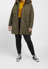 Chaqueta parka verde oliva con capucha de piel sintética, cierre de cremallera, cintura ajustada y bolsillos delanteros. Llevada sobre una camiseta mostaza y pantalones vaqueros negros desgastados.