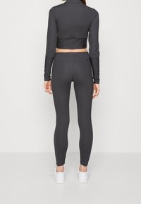 Kvinna i mörkgrå figurnära leggings och matchande kortärmad topp med lång ärm, stående med ryggen mot kameran framför en enfärgad bakgrund.