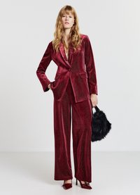 Samtenes roter Anzug mit einem strukturierten Blazer und weit geschnittenen Hosen, mit einem dezenten Diamantmuster. Eine schwarze, flauschige Handtasche ergänzt den Look.