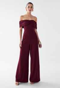 Burgundijos spalvos off-the-shoulder kombinezonas su raukiniais, plačiomis kelnėmis ir prigludusia liemene; pagamintas iš lygios medžiagos su minimaliu išbaigtumu.