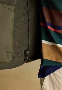 Oliwkowa polarowa kurtka z teksturowanej tkaniny i regulowanym sznurkiem. Zestawiona z wielokolorową koszulą w paski w odcieniach zieleni, brązu i niebieskiego.