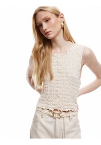 Top texturizado beige claro con volantes verticales y un escote con forma de pico, combinado con pantalones de color claro que llevan un cinturón de hebilla de tono dorado.