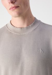 Lys grå hettegenser med ribbestrikket halslinning og subtilt preget logo på brystet. Glatt stoff med en lett børstet tekstur.
