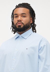 Hombre con barba y rastas, vistiendo una camisa de botones azul claro con un patrón y un pequeño logo rojo, blanco y azul en el pecho.