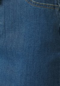 Close-up van blauwe denimstof met zichtbare diagonale geweefde textuur en gele stiksels nabij de rand van de zak.