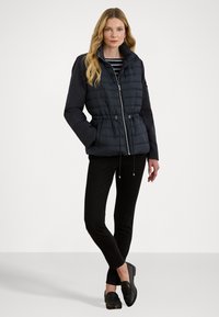Pufferjack in donkerblauw met een gequilt textuur, elastische trekkoord taille, ritssluiting aan de voorkant en zijzakken; gecombineerd met zwarte broek en loafers.