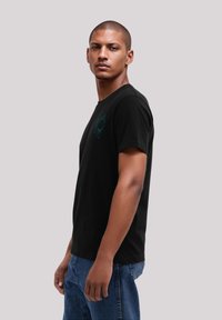 Schwarzes Baumwoll-T-Shirt mit kurzen Ärmeln, das ein türkisfarbenes Sonnenmotiv auf der linken Brust zeigt. Das Model posiert seitlich und trägt eine blaue Jeans.