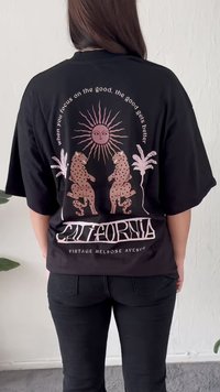 Persona que lleva una camiseta negra con diseño rosa de un sol, dos leopardos manchados enfrentados, palmeras y el texto "California Vintage Melrose Avenue."