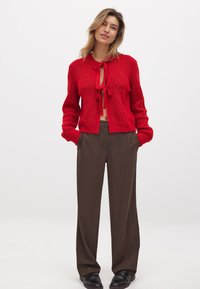 Roter gestrickter Cardigan mit einem verkürzten Design, gebundenem Vorderteil, langen Ärmeln, kombiniert mit braunen weiten Hosen und schwarzen Schuhen.