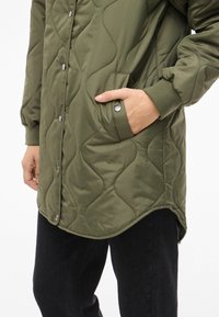 Grön quiltad jacka med slät yta, tryckknappar och sidosfickor; har en rundad fåll och ribbade ärmslut.