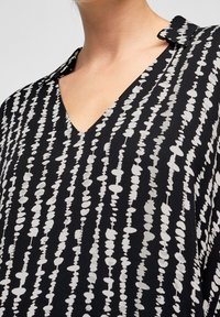 Svart blus med V-ringning, prydd med ett grått abstrakt mönster av avlånga former och ränder, tillverkad av ett lätt material.