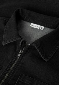 Veste en denim noir avec un col, fermeture éclair sur le devant et une surface texturée. Étiquette visible à l'intérieur du col. Deux poches à la poitrine.