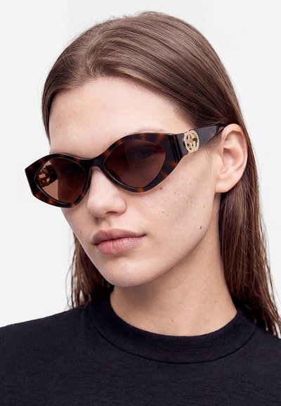 Ung kvinna med rakt brunt hår som bär solglasögon i cat-eye-stil med sköldpaddsmönster och gulddetalj på loggan samt en svart topp mot en enfärgad bakgrund.