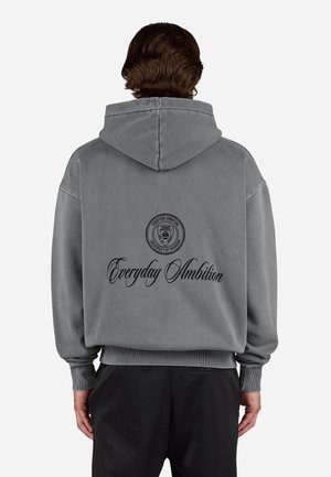 Person mit kurzen braunen Haaren, die einen grauen Kapuzenpullover mit dem Text "Everyday Ambition" und einem universitätsähnlichen Emblem auf dem Rücken trägt, steht still.