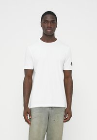 Biele tričko s krátkym rukávom a okrúhlym výstrihom, vyrobené z hladkej tkaniny, s malým čiernym logom na ľavom rukáve.