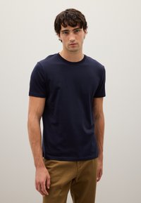 Giovane uomo in piedi contro un muro semplice, indossa una maglietta a maniche corte blu navy e pantaloni marrone chiaro, guarda direttamente davanti a sé.