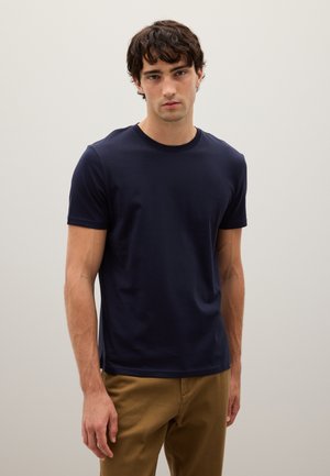Young man standing against plain wall, wearing a navy blue short-sleeve t-shirt and light brown pants, looking directly ahead.