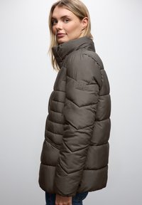 Ciemnooliwkowa puchowa kurtka z wysokim kołnierzem, pikowaną teksturą i naszywką z logo na lewym rękawie. Ma zamykane na zamek kieszenie po bokach.